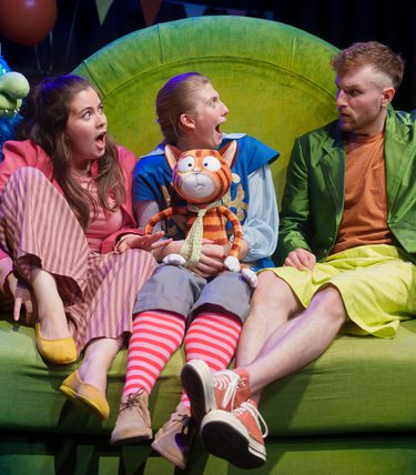 Three performers sit on a large bright green sofa, reacting with surprise while holding a puppet of a wide-eyed orange cat. Colourful monster puppets peek over the sides of the sofa in this playful children’s theatre scene.