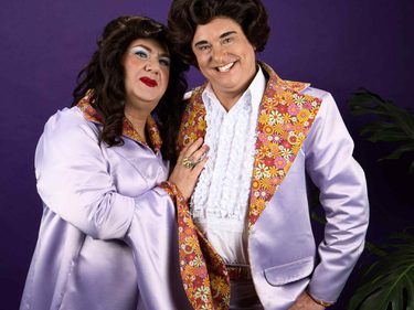 Two performers dressed as the double act Pearl & Dean stand against a purple backdrop, wearing matching lilac satin outfits with colourful floral-patterned lapels and cuffs. One performer rests a hand on the other's chest, both smiling warmly at the camera.